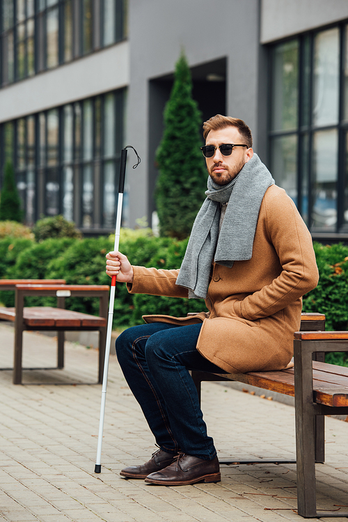 Blind man in sunglasses holding walking stick while sitting on bench