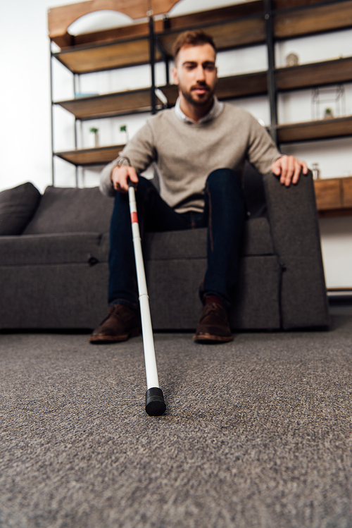 Selective focus of visually impaired man holding walking stick while sitting on sofa in living room