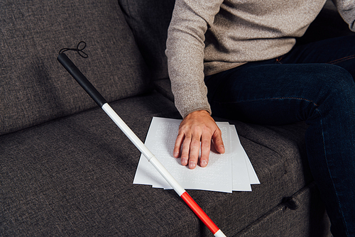 Cropped view of blind man reading braille font beside walking stick on sofa