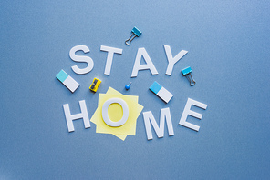 Top view of stay home lettering near sticky notes, binder clips and pencil sharpener on blue surface