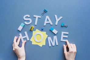 Top view of woman touching stay home lettering near erasers, binder clips and sticky notes on blue surface