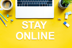 Top view of stay online lettering near laptop, coffee and stationery on yellow background