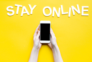 Cropped view of woman holding smartphone with blank screen and lettering stay online on yellow surface