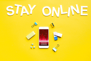 Top view of smartphone with trading courses app, office supplies and stay online lettering on yellow surface