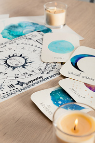 Selective focus of watercolor drawings with moon phases on cards and zodiac signs on wooden table with candles