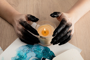 Cropped view of witch with black paint on hands holding candle by cards with watercolor zodiac signs on table