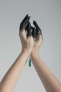 Cropped view of witch with black paint on hands holding green crystal on chain isolated on grey