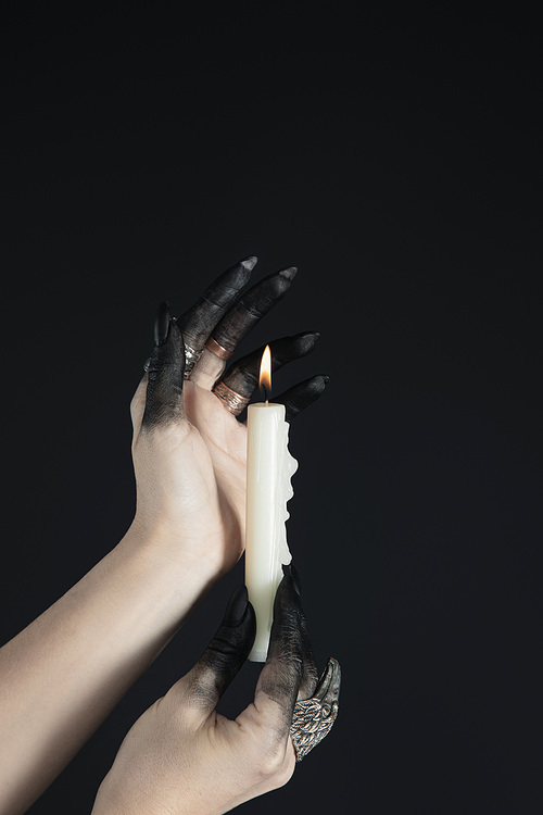 Cropped view of black painted witch hands holding burning candle isolated on black