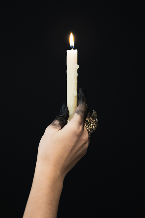 Cropped view of witch with black paint on hand and jewelry ring holding candle isolated on black