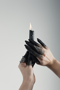Cropped view of witch hands with black painted fingers and jewelry rings holding candle isolated on grey