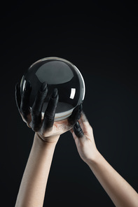 Cropped view of black painted hands of witch holding transparent crystal ball isolated on black