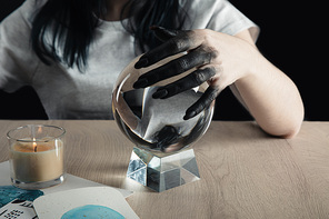 Cropped view of magician with black painted hand holding crystal ball by watercolor paintings and candle on table isolated on black