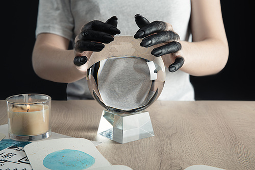 Cropped view of witch with black dyed hands holding crystal ball by candle and watercolor drawings on table isolated on black