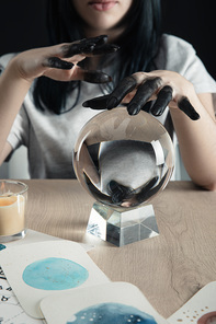 Cropped view of magician with black paint on hands, crystal ball and watercolor paintings on table isolated on black