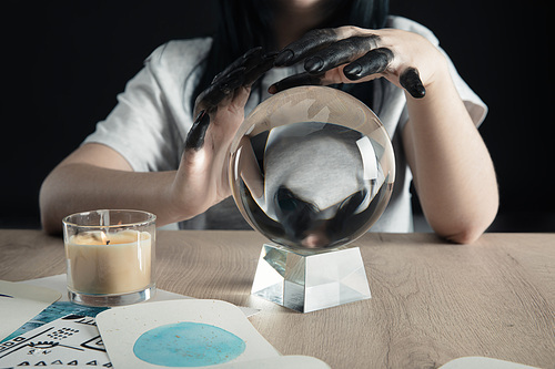 Cropped view of witch with black painted hands, crystal ball and watercolor zodiac signs on cards on table isolated on black