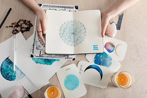 Top view of astrologer holding notebook with watercolor drawings and zodiac signs on cards on table