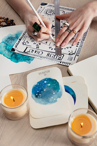 Selective focus of fortuneteller drawing birth chart by cards with watercolor paintings and candles on table