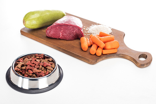 dry pet food in bowl near raw meat, , carrot and zucchini on wooden cutting board isolated on white