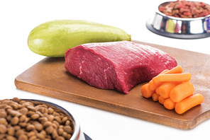 selective focus of dry pet food in bowls near raw meat, carrot and zucchini on wooden cutting board isolated on white