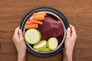 cropped view of woman holding pet bowl with raw meat and vegetables on wooden background