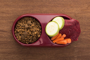 top view of dry pet food and raw meat with vegetables in bowl on wooden table