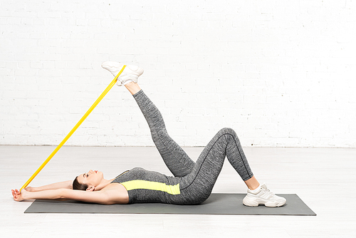 side view of woman in sportswear working out with resistance band while lying on fitness mat near white brick wall