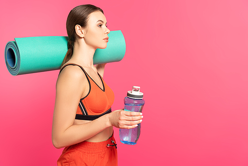 attractive sportswoman holding sports bottle with water and fitness mat isolated on pink