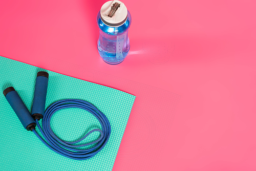 top view of sports bottle with fresh water near skipping rope on fitness mat on pink