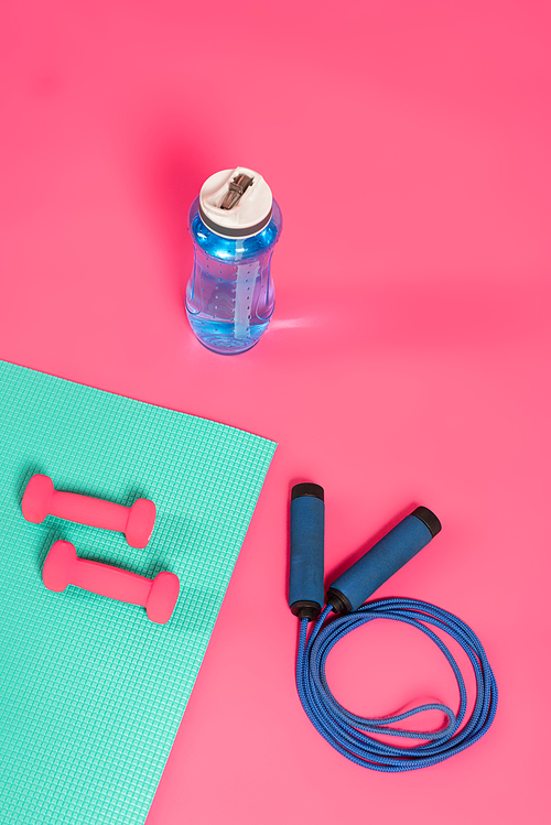 top view of sports bottle with fresh water near skipping rope and dumbbells on fitness mat on pink