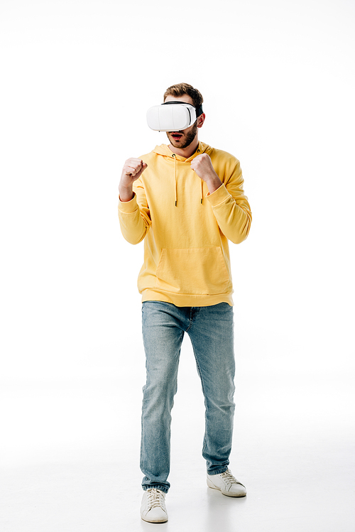 young man in blue jeans and yellow hoodie imitating boxing while using virtual reality headset on white background