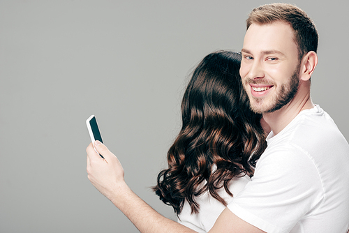 handsome smiling man embracing woman and using smartphone isolated on grey