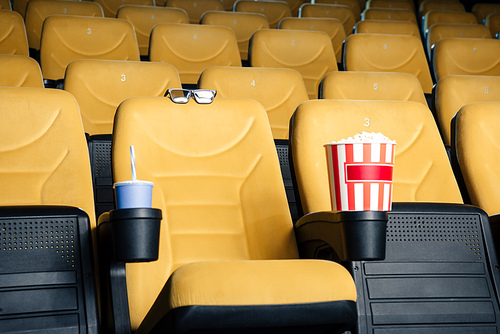 orange cinema seats with paper cups of soda and popcorn in cup holders, and 3d glasses