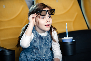 cute surprised child watching movie in cinema while sitting near paper cup