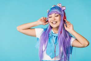 Smiling anime girl in purple wig listening music in headphones isolated on blue