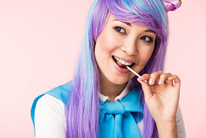 Happy asian otaku girl in wig eating lollipop isolated on pink