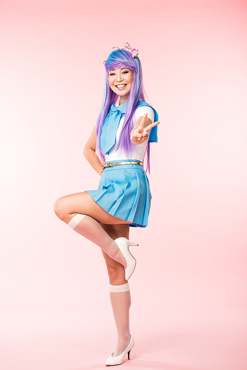 Full length view of happy asian anime girl standing on one leg and showing peace sign on pink