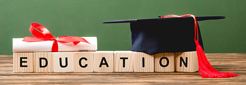 panoramic shot of wooden blocks with letters, diploma and academic cap on wooden surface isolated on green