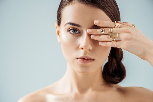 young nude woman with shiny makeup and golden rings hiding eye behind hand isolated on grey