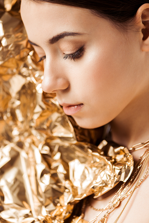 young woman with shiny makeup and golden foil in necklaces posing with closed eyes