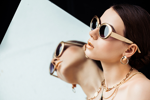 young naked woman in sunglasses, golden necklaces holding mirror isolated on black