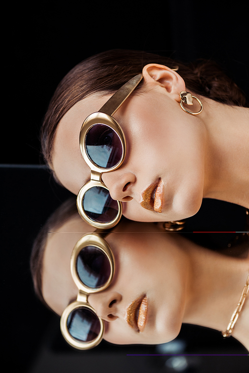 young woman in sunglasses, golden jewelry lying on mirror isolated on black
