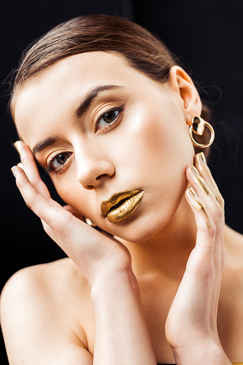 young naked woman with golden makeup and golden paint on fingers touching face isolated on black