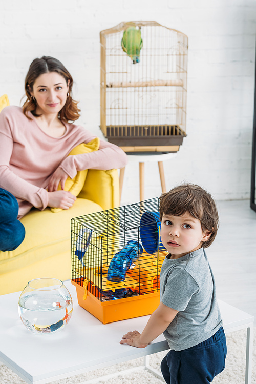 selective focus of cute child standing near table with fish bowl and pet cage, and smiling mother resting on yellow sofa