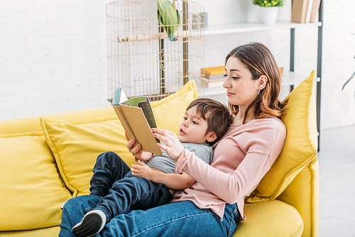 smiling woman with cute child reading book on sofa together