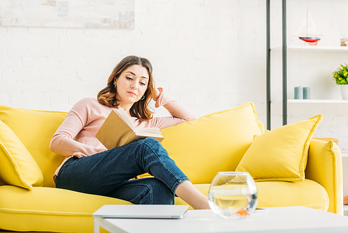 beautiful young woman reading book on sofa near table with fish bowl