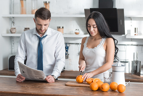 attractive brunette girl cutting orange on cutting board near man reading newspaper