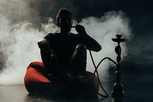 silhouette of man sitting on bean bag chair and smoking hookah in darkness