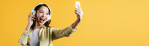 happy asian girl taking selfie on smartphone, singing and listening music with headphones, isolated on yellow