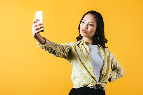 attractive asian girl taking selfie on smartphone isolated on yellow