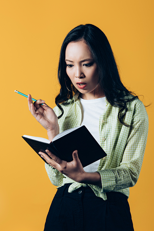 shocked asian girl writing in notebook with pen, isolated on yellow